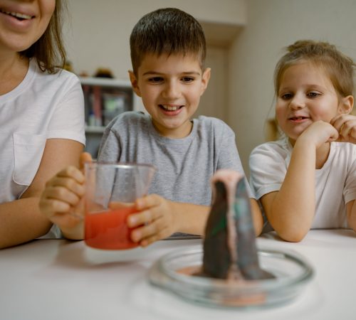 Mom and children at home are conducting an experiment with volcanic eruption. Boy pours liquid into volcanic and rejoices at reaction of eruption. Experiments at home, fun physics with kids